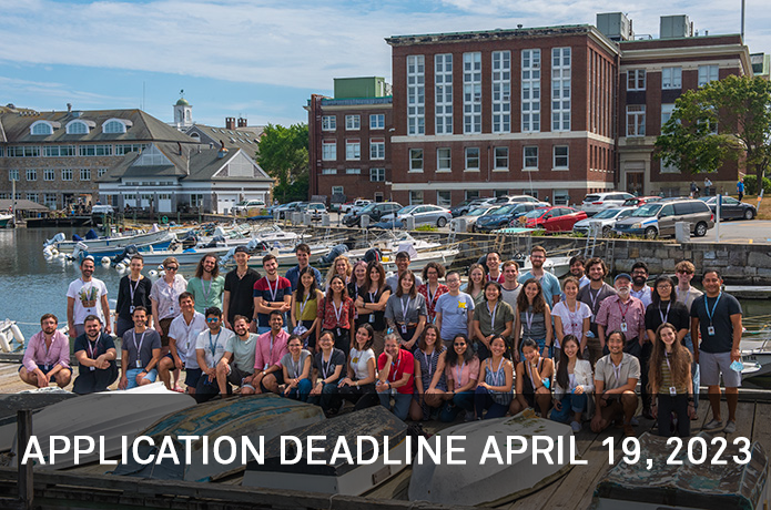 group photo of student on a dock with upside down rowboats in front of them and a brick building and pond behind them